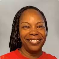 Smiling woman with braided hair, wearing a red shirt and hoop earrings, posing in front of a plain light background.